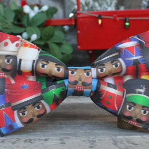 May include: A festive bow tie featuring a colorful pattern of nutcracker soldiers. The bow tie is made of fabric and has a red, green, blue, and white color scheme.