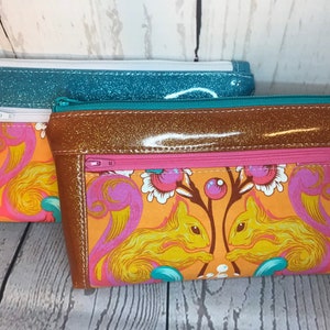 May include: Two zippered pouches with different patterns. The top pouch is teal blue with white glitter. The bottom pouch is brown with gold glitter and features a colorful squirrel print.