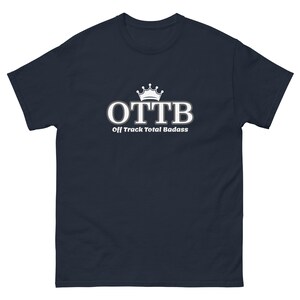 May include: A navy blue short-sleeve t-shirt featuring a white graphic on the chest. The graphic displays a crown icon above the large white text "OTTB" and the smaller white text "Off Track Total Badass" centered below the main lettering.