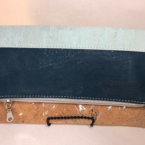 May include: A cork clutch with a zipper closure. The clutch is made of three different colors of cork: light blue, dark blue, and brown. The clutch has a silver zipper and a silver chain strap.