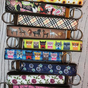 May include: A set of ten keychains with different colorful patterns. The keychains are made of fabric and have a brown leather strap. The patterns include sunflowers, paisley, plaid, horses, cats wearing glasses, dogs wearing glasses, flowers, and camouflage.