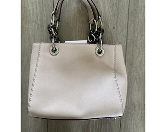 May include: A light pink handbag with silver-tone hardware and chain handles. The bag has a structured shape with a top handle and a shoulder strap. The chain handles are a mix of silver and brown links. The bag is sitting on a wooden surface.