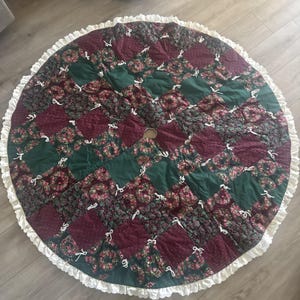 May include: A round Christmas tree skirt with a patchwork design. The skirt features alternating squares of dark red floral fabric and solid green fabric. White bows are sewn on the seams. The edge is trimmed with a white ruffled border.