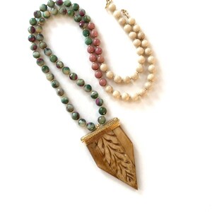 May include: A long necklace with green, pink, and cream colored beads and a carved wooden pendant with a gold trim.