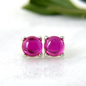 May include: A pair of stud earrings with vibrant magenta-colored, rounded gemstones. The gems are set in a silver-colored metal with a four-prong setting. The earrings are displayed on a white surface with a blurred green background.