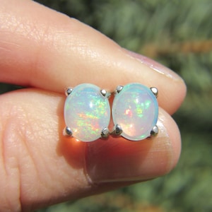 May include: A pair of oval-shaped opal stud earrings. The opals have a milky white base color with flashes of iridescent colors. The earrings are set in a silver-colored metal.