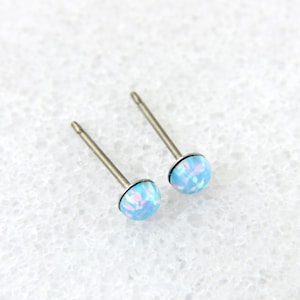 May include: A pair of stud earrings with light blue opal-like stones. The earrings have a silver-colored post and a round, domed stone. The stones have a mix of blue and white colors. The earrings are set against a white background.
