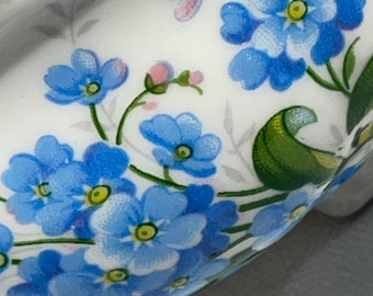 May include: Close-up of a white ceramic object with a gold rim, adorned with a floral pattern. The design features clusters of light blue flowers with yellow centers, green leaves, and pink buds. The background is white with subtle gray leaf accents.
