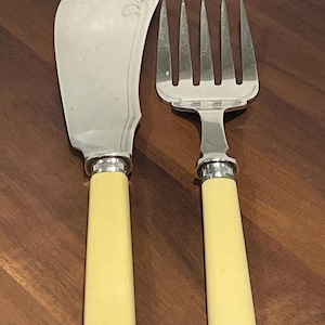 Puede incluir: Un juego de dos cubiertos de servir plateados con mangos de color marfil. El cuchillo tiene una hoja curva y el tenedor tiene cinco dientes.