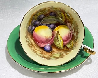 May include: A vintage teacup and saucer set with a green saucer and a cream-colored teacup with a floral design featuring peaches and grapes.