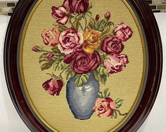 May include: Oval framed needlepoint art featuring a bouquet of roses in shades of red, pink, and yellow, arranged in a blue vase. The background is a light tan color, and the frame is dark brown with a gold inner border.