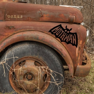 Puede incluir: Un camión Dodge vintage oxidado con una calcomanía de murciélago negro en el guardabarros. La calcomanía dice "Mothman".