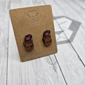 May include: A pair of wooden earrings featuring a cartoon character with a green face, orange hair, and a black hat. The earrings are on a brown card with the text "Brilliant Creations" and "@brilliantcreations".