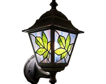May include: A black outdoor wall-mounted lantern with a stained glass design. The glass panels feature green leaf patterns against a blue and white background. The lantern has a decorative finial and a curved arm for mounting.