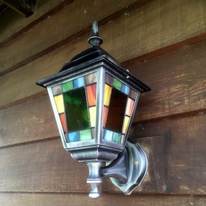 May include: A decorative outdoor wall-mounted lantern with a stained glass design. The lantern features a silver-colored metal frame and a multi-colored glass panel design with green, blue, yellow, orange, and brown hues. The lantern is mounted on a wooden surface.
