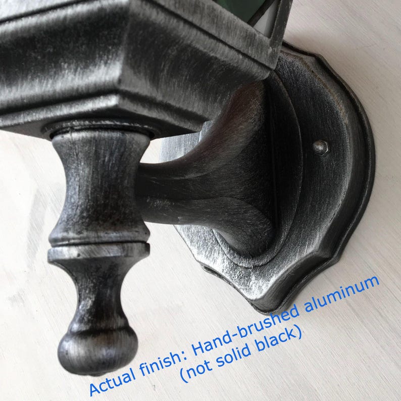May include: Close-up of a hand-brushed aluminum outdoor wall light fixture. The fixture has a decorative, ornate design with a dark gray finish and a silver brushed effect. The text in the image reads: Actual finish: Hand-brushed aluminum (not solid black).