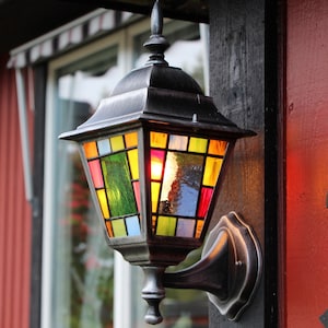 May include: A decorative outdoor wall-mounted lantern with a stained glass design. The lantern features a dark metal frame and a colorful array of glass panels in shades of red, yellow, green, and blue. The lantern is mounted on a dark wooden post.