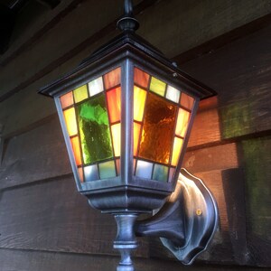 May include: A decorative outdoor wall-mounted lantern with a stained glass design. The lantern features a dark metal frame and colorful glass panels in shades of green, orange, yellow, and red. The lantern is mounted on a wooden wall.