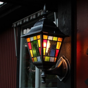 May include: A decorative outdoor lantern with a stained glass design. The lantern features a black metal frame and a colorful array of glass panels in shades of red, yellow, green, and blue. It is mounted on a dark wooden post.