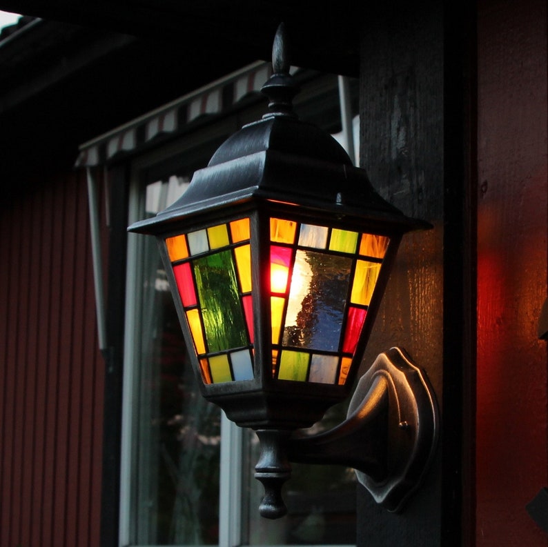 May include: A decorative outdoor lantern with a stained glass design. The lantern features a black metal frame and a colorful array of glass panels in shades of red, yellow, green, and blue. It is mounted on a dark wooden post.