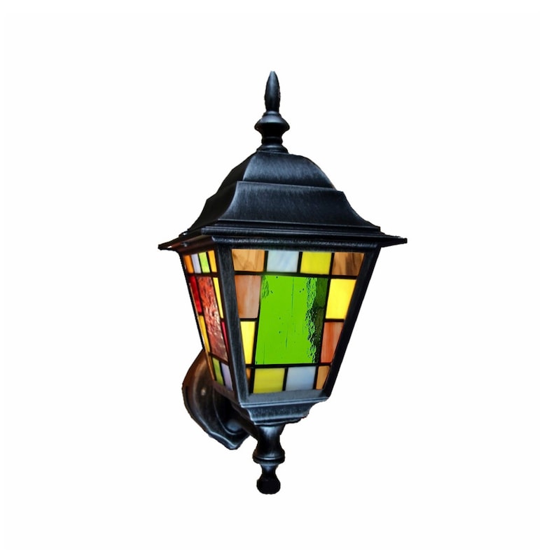 May include: Black outdoor wall-mounted lantern with a stained glass design. The lantern features a multi-colored glass panel with red, yellow, green, and brown hues. The metal frame has a decorative top and bottom, and is mounted on a wall bracket.
