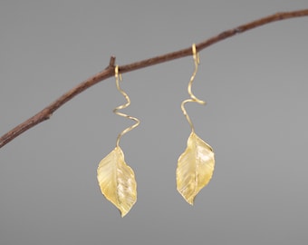 May include: A pair of gold-colored leaf earrings. Each earring features a detailed leaf pendant suspended from a curved, spiral-shaped wire. The earrings are displayed against a neutral gray background, with a brown twig in the upper left.