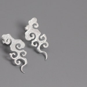 Könnte beinhalten: Ein Paar silberfarbene Ohrringe in Wolkenform. Jeder Ohrring hat ein wirbelndes, strukturiertes Design mit einem Steckerverschluss. Die Ohrringe werden vor einem grauen Hintergrund gezeigt.