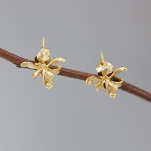 Könnte beinhalten: Ein Paar goldfarbene Ohrstecker, jeweils in Form einer zarten Blume. Die Ohrringe werden auf einem dünnen, braunen Ast vor einem weichen, grauen Hintergrund präsentiert. Das florale Design hat detaillierte Blütenblätter.