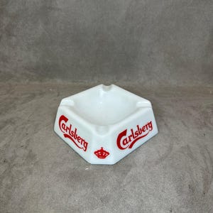 Puede incluir: Cenicero de cerámica blanca con un logotipo rojo de Carlsberg a cada lado. El logotipo incluye una corona.
