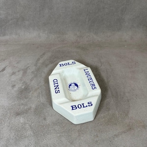 May include: White ceramic ashtray with blue lettering. The ashtray is divided into three sections labeled "GINS", "LIQUEURS", and "BOLS".