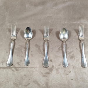 May include: Five pieces of silverware, including forks and spoons, are arranged on a neutral-toned surface. The silverware has a shiny, metallic finish, reflecting the light. The forks have four tines, and the spoons have a rounded bowl.