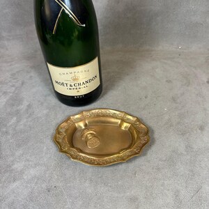May include: A gold-colored metal tray with a decorative grapevine design. The tray has a raised edge and a small, round indentation in the center. A bottle of Moët & Chandon Imperial Brut champagne is in the background.