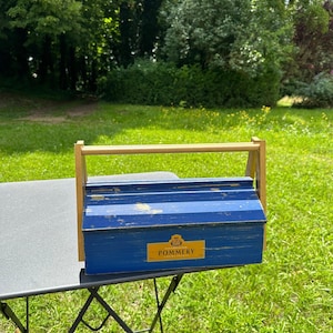 Peut inclure: Une boîte à outils en bois bleu avec une poignée dorée et une étiquette dorée indiquant "Champagne Pommery". La boîte à outils est posée sur une table pliante noire.