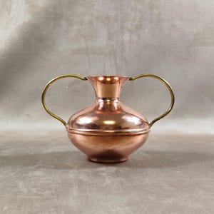 May include: A copper-colored vase with two brass handles. The vase has a rounded body and a flared neck. The handles are curved and attached to the top of the vase. The vase is made of metal and has a shiny finish.