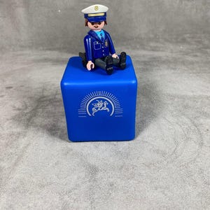 以下が含まれることがあります： 青いプラスチック製の警察官のおもちゃのフィギュアが、白いロゴの付いた青い立方体の上に座っています。ロゴには馬と星が描かれています。
