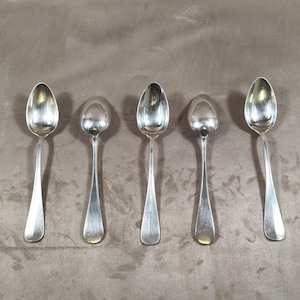 May include: Five silver spoons arranged in a row. Each spoon has a rounded bowl and a long, slender handle. The spoons are made of a shiny, reflective metal, likely silver or a silver-colored alloy. The spoons are of varying sizes.