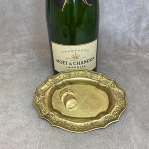 May include: A gold-colored metal tray with an ornate design featuring grapes and vines. A champagne cork with the Moët & Chandon logo is sitting in the tray.