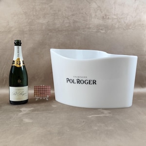 Op de afbeelding: Een fles Pol Roger champagne naast een witte champagne-emmer met het Pol Roger-logo. Een kleine visitekaartje staat voor de emmer. De champagnefles is groen met een gouden etiket.