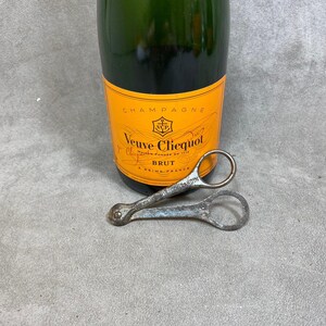 Puede incluir: Una botella de champán Veuve Clicquot Brut con un abridor de botellas de plata. La botella es naranja y verde con el texto "Champagne Veuve Clicquot Ponsardin fondée en 1772 Brut Reims France".