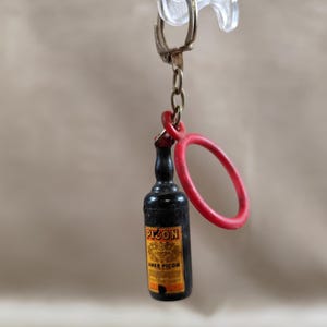 Peut inclure: Un porte-clés miniature en forme de bouteille noire avec un anneau rouge et une chaîne dorée. La bouteille porte une étiquette jaune avec le texte "Picon" et "Amer Picon". Le porte-clés est attaché à un fermoir en plastique transparent.