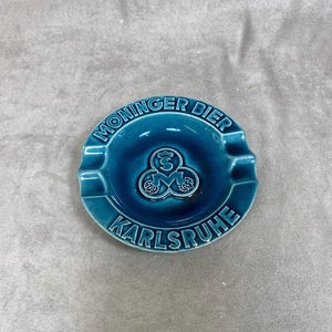 May include: A blue ceramic ashtray with the words "Moninger Bier Karlsruhe" and a logo of three hops with a crown on top.