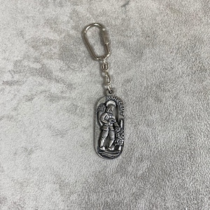 May include: Silver keyring featuring a detailed relief of a man in traditional clothing standing in front of a grapevine. The keyring has a silver loop for attaching to keys.