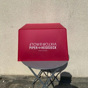 Peut inclure: Une boîte rectangulaire rouge avec le texte "Piper Heidsieck Viktor & Rolf Rose Sauvage" imprimé dessus. La boîte est posée sur une table pliante en métal noir.