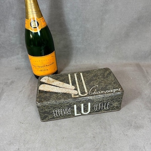 Peut inclure: Une boîte en métal vintage verte et dorée avec les mots "Lefevre LU Utile" et "Champagne" imprimés dessus. La boîte présente un motif de flûte à champagne et une bouteille de champagne.