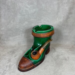 May include: A vintage ceramic ashtray shaped like a green boot with a brown toe and trim. A brown and gold pipe is attached to the side. The top of the boot has a circular ashtray.