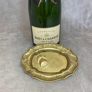 May include: A gold-colored metal champagne bottle stopper tray with a grapevine design. The tray has a small, round indentation in the center, where a champagne cork is resting. The cork is silver and has the words "Moët & Chandon" printed on it.