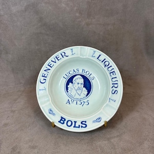 May include: A white ceramic ashtray with a blue and white design. The design features a portrait of Lucas Bols, the founder of the Bols liqueur company, and the text "Lucas Bols A° 1575" and "Genever & Liqueurs Bols".