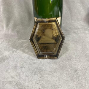 May include: A hexagonal glass ashtray with a raised design of a person's profile. The ashtray is brown and has gold lettering around the edge.