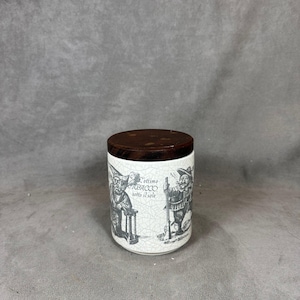 May include: White ceramic jar with a brown wooden lid. The jar has a black and white illustration of two men with the text "l'ottimo tabacco sotto il sole".