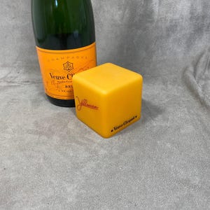 以下が含まれることがあります： 黄色のヴーヴ・クリコシャンパンボトルストッパーで、底にブランド名が印刷されています。ストッパーは立方体の形をしています。